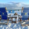 A cozy beach bonfire setup at sunset featuring six chairs arranged around a fire pit, illuminated by tiki torches, with a large Bluetooth speaker playing music, and a s’mores kit ready for roasting.​